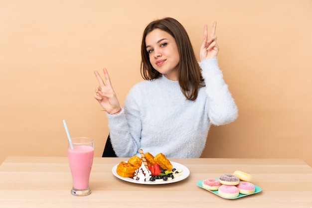 Teenager girl eating waffles isolated on beige wall showing victory sign with both hands