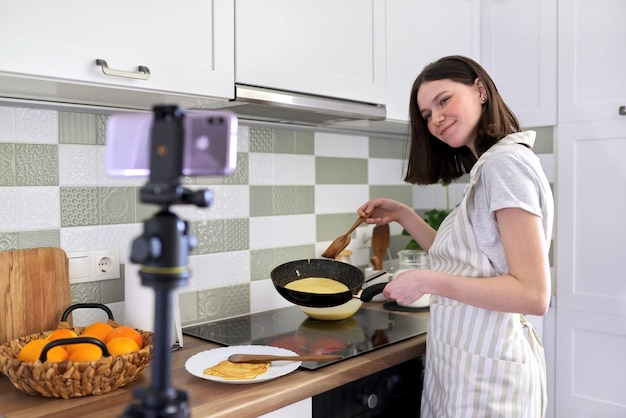 Teenager girl, food blogger cooking pancakes with orange at home in kitchen, filming video recipe. Female with frying pan near stove. Hobbies, video channel with followers, children and teenagers