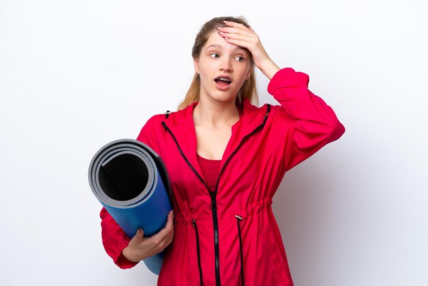 Teenager girl going to yoga classes while holding a mat isolated on white bakcground doing surprise gesture while looking to the side