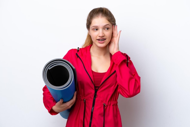Teenager girl going to yoga classes while holding a mat isolated on white bakcground listening to something by putting hand on the ear