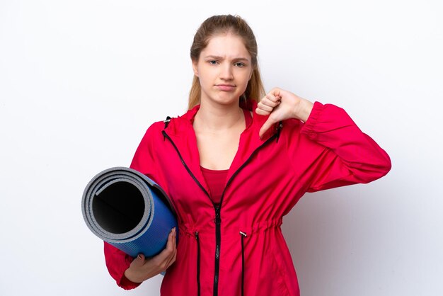 Teenager girl going to yoga classes while holding a mat isolated on white bakcground showing thumb down with negative expression