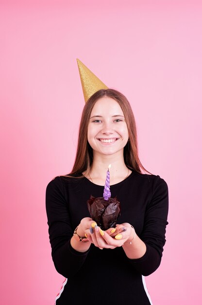 Ragazza dell'adolescente nel compleanno dorato che tiene focaccina con candele, esprimendo un desiderio su sfondo rosa