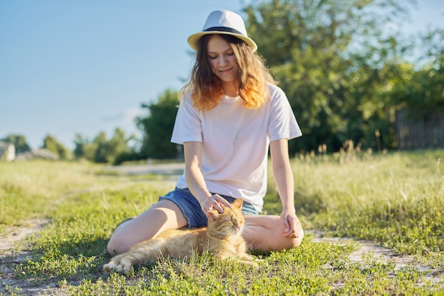 Teenager girl in hat on nature playing with red cat, rustic style