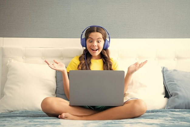 Teenager girl in headphones relax on bed at home using laptop Child in earphones browse internet on computer Expressive emotional excited teen girl