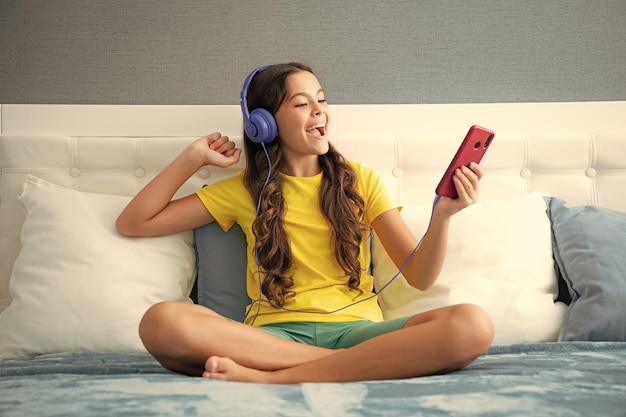 Teenager girl in headphones relax on bed at home using phone Child in earphones browse internet on smartphone Expressive emotional excited teen girl