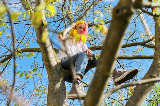 Девушка-подросток, хипстер, сидящая высоко на дереве. Весна, природа фон неба. Подростковый возраст, характер, образ жизни, поведение подростков