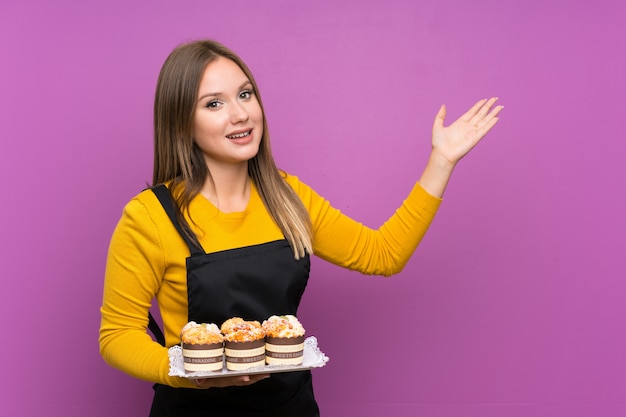 Teenager girl holding lots of different mini cakes over isolated purple background extending hands to the side for inviting to come