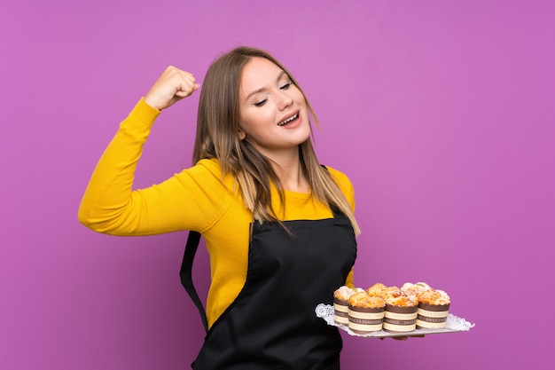 Teenager girl holding lots of different mini cakes over isolated purple celebrating a victory