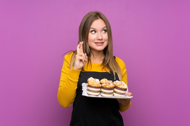 Девушка-подросток держит много различных мини-пирожных над фиолетовой стеной с скрещиванием пальцев