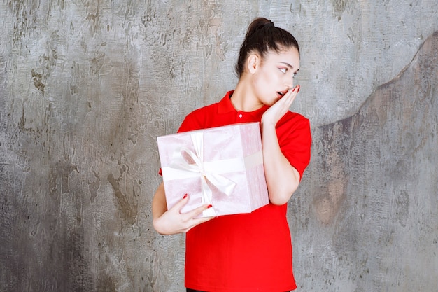 Teenager girl holding a pink gift box wrapped with white ribbon and looks thoughtful. 