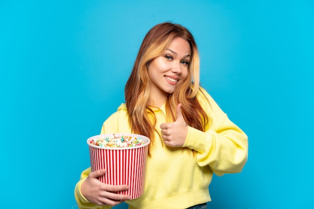 Teenager girl holding popcorns over isolated blue background giving a thumbs up gesture