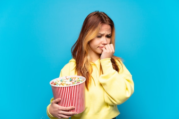 Девушка-подросток держит попкорн на изолированном синем фоне, сомневаясь
