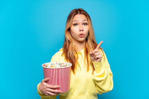 Teenager girl holding popcorns over isolated blue background intending to realizes the solution while lifting a finger up