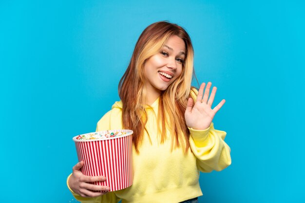 Девушка-подросток держит попкорн на изолированном синем фоне, салютуя рукой со счастливым выражением лица