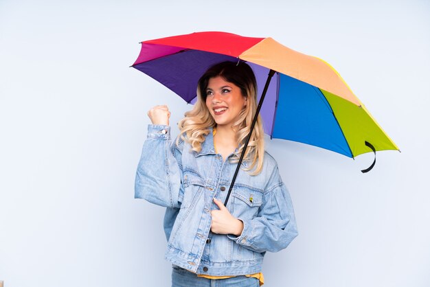 Teenager girl holding an umbrella on blue wall celebrating a victory