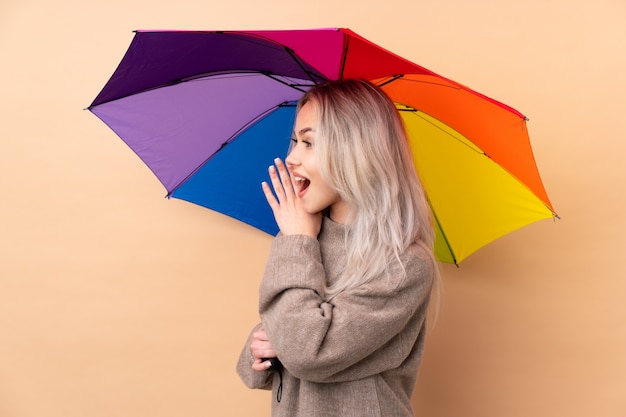 Teenager girl holding an umbrella over wall shouting with mouth wide open