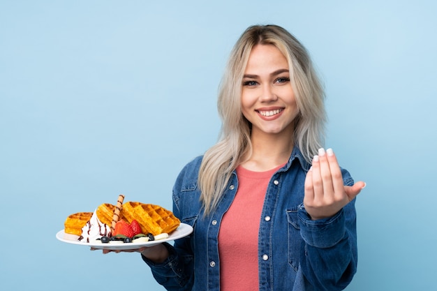 Teenager girl holding waffles over blue wall inviting to come with hand. Happy that you came