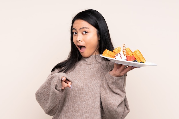 Teenager girl holding waffles isolated on beige surprised and pointing front