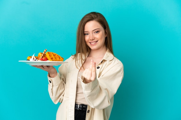 Teenager girl holding waffles over isolated blue background making money gesture