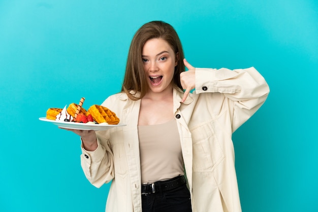 Teenager girl holding waffles over isolated blue background making phone gesture. Call me back sign