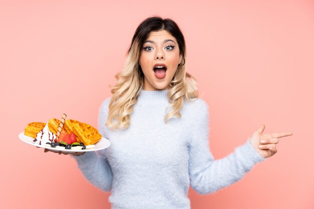 Teenager girl holding waffles on isolated pink wall surprised and pointing side