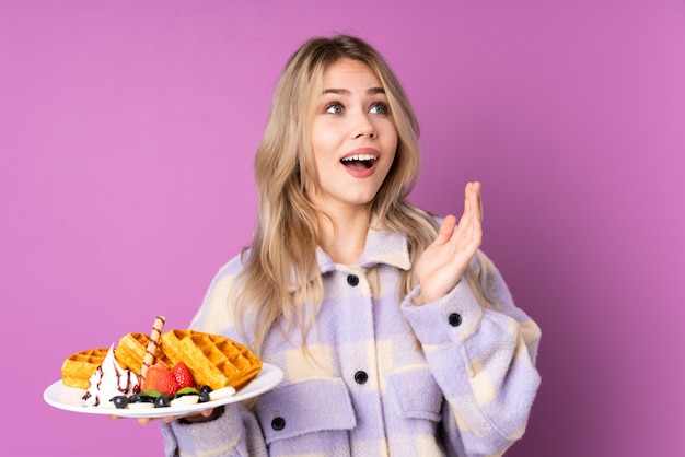 Photo teenager girl holding waffles isolated on purple wall with surprise facial expression