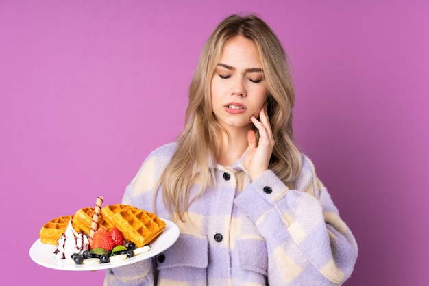 Teenager girl holding waffles isolated on purple wall with toothache