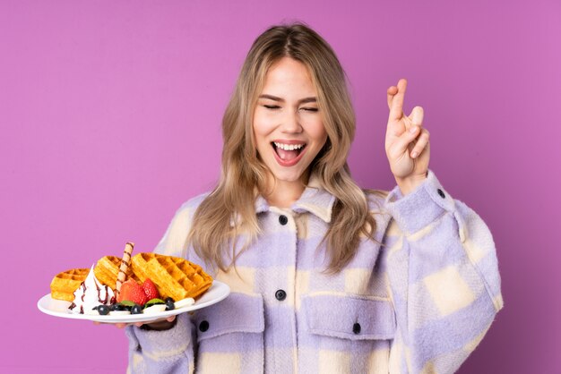 Девочка-подросток держит вафли на фиолетовой стене со скрещенными пальцами
