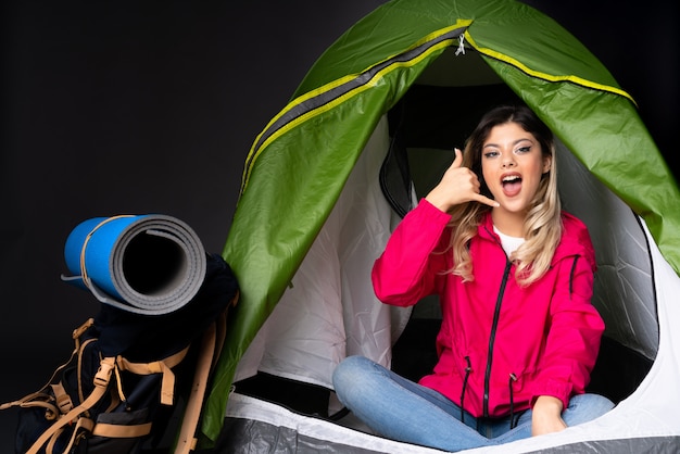 Photo teenager girl inside a camping green tent isolated on black wall making phone gesture and doubting