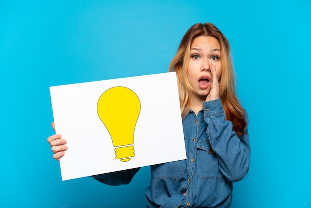 Teenager girl over isolated blue background holding a placard with bulb icon and shouting