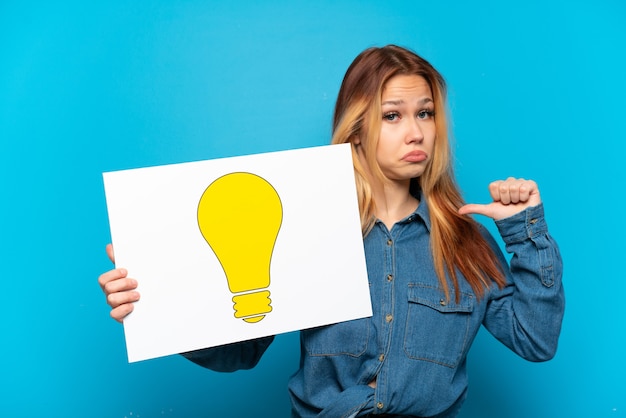 Teenager girl over isolated blue background holding a placard with bulb icon with proud gesture