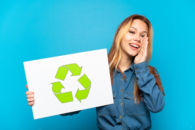 Teenager girl over isolated blue background holding a placard with recycle icon and shouting