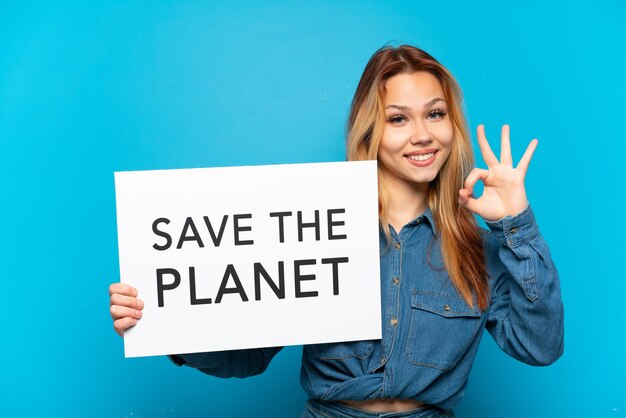Девушка-подросток на изолированном синем фоне держит плакат с текстом «Спасите планету» и празднует победу
