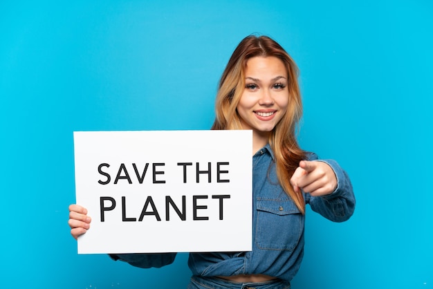 Teenager girl over isolated blue background holding a placard with text Save the Planet and pointing to the front