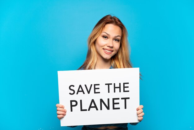 Девушка-подросток на изолированном синем фоне держит плакат с текстом «Спасите планету» со счастливым выражением лица