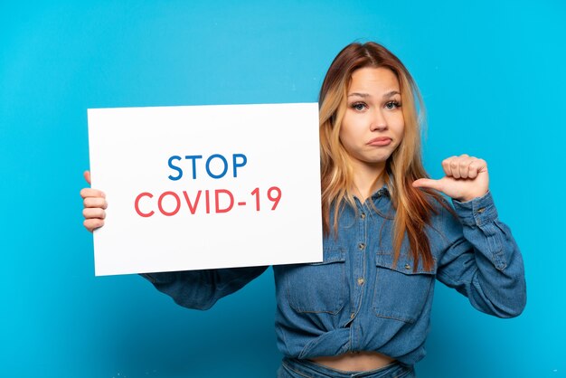 Teenager girl over isolated blue background holding a placard with text Stop Covid 19 with proud gesture