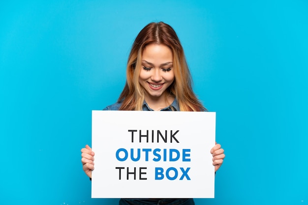 Teenager girl over isolated blue background holding a placard with text Think Outside The Box