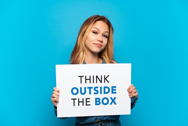 Teenager girl over isolated blue background holding a placard with text Think Outside The Box