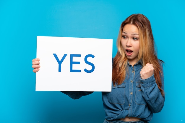 Teenager girl over isolated blue background holding a placard with text yes and celebration a victory