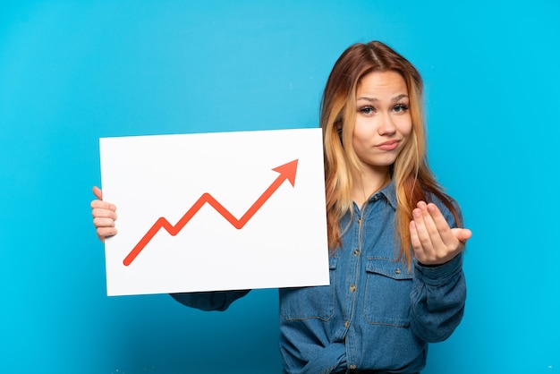 Teenager girl over isolated blue background holding a sign with a growing statistics arrow symbol and doing coming gesture