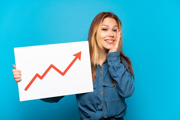 Teenager girl over isolated blue background holding a sign with a growing statistics arrow symbol and shouting