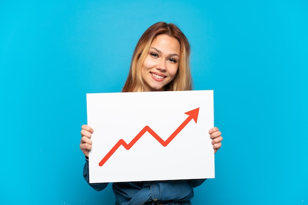 Teenager girl over isolated blue background holding a sign with a growing statistics arrow symbol with happy expression