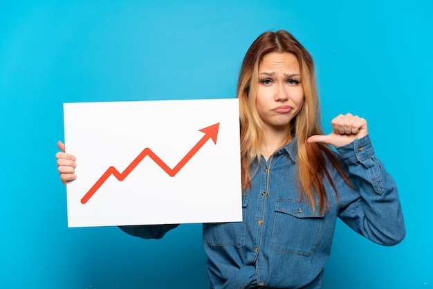 Foto ragazza dell'adolescente sopra fondo blu isolato che tiene un segno con un simbolo della freccia di statistiche in crescita con gesto orgoglioso