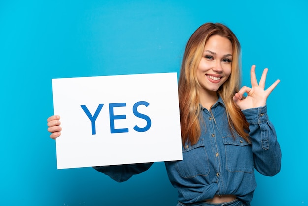 Teenager girl over isolated blue surface holding a placard with text YES