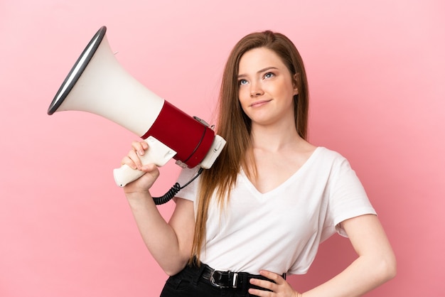 Ragazza dell'adolescente sopra fondo rosa isolato che tiene un megafono e che pensa