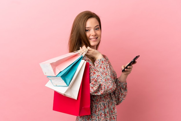 Teenager girl over isolated pink background holding shopping bags and writing a message with her cell phone to a friend