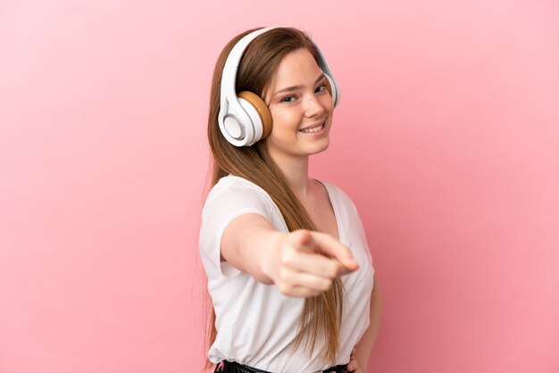 Девушка-подросток на изолированном розовом фоне, слушая музыку и указывая на фронт