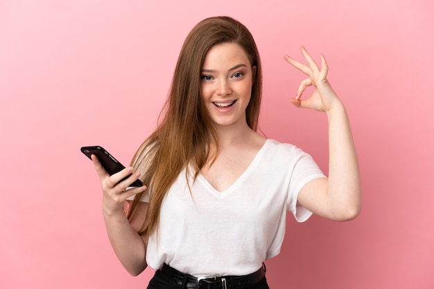 Teenager girl over isolated pink background using mobile phone and doing OK sign