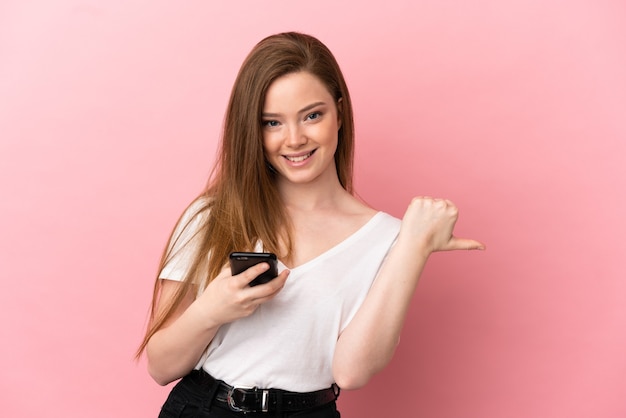 Teenager girl over isolated pink background using mobile phone and pointing to the lateral