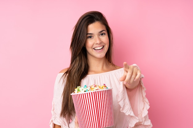 Teenager girl over isolated pink wall holding a big bucket of popcorns while pointing front